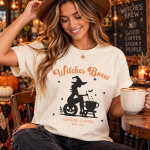 May include: A cream-coloured t-shirt featuring a graphic print with the text "Witches Brew" in orange, a black silhouette of a witch sitting on a pumpkin stirring a cauldron, and the text "COFFEE COVEN EST. 1604" in orange at the bottom.