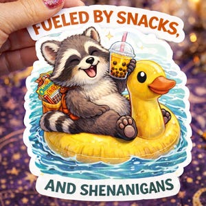 May include: A sticker featuring a cartoon raccoon floating on a yellow inflatable duck in water. The raccoon holds a bubble tea and wears a backpack with snacks. Text reads "Fueled by Snacks, and Shenanigans."