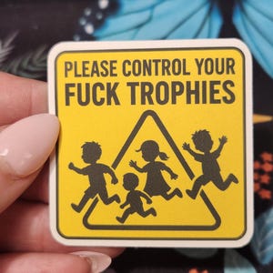 Op de afbeelding: Een gele vierkante sticker met afgeronde hoeken. De sticker bevat de tekst "PLEASE CONTROL YOUR FUCK TROPHIES" boven een zwarte driehoek met rennende stokfiguren. De sticker wordt door een persoon vastgehouden.