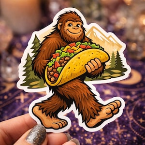 May include: A sticker featuring a cartoon Bigfoot carrying a large taco. The Bigfoot is brown and furry, with a smiling face. The taco is filled with lettuce, tomatoes, and meat. The background includes mountains and pine trees.