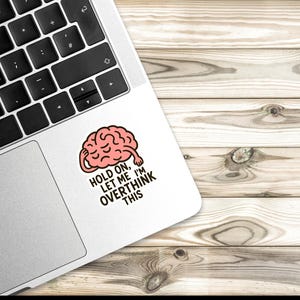 May include: A white sticker with a cartoon brain graphic on a laptop. The brain is pink with a red outline, and the sticker has the text "HOLD ON, LET ME I'M OVERTHINK THIS" in black. The laptop is on a wooden surface.