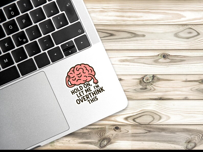 May include: A white sticker with a cartoon brain graphic on a laptop. The brain is pink with a red outline, and the sticker has the text "HOLD ON, LET ME I'M OVERTHINK THIS" in black. The laptop is on a wooden surface.