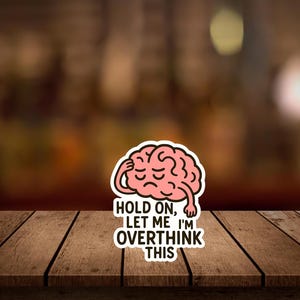 May include: A sticker featuring a pink brain with a sad expression, touching its head. The sticker has the text "HOLD ON, LET ME I'M OVERTHINK THIS" in black and white, set against a blurred background.