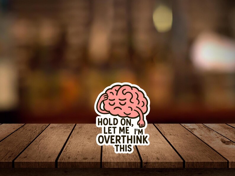 May include: A sticker featuring a pink brain with a sad expression, touching its head. The sticker has the text "HOLD ON, LET ME I'M OVERTHINK THIS" in black and white, set against a blurred background.