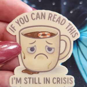以下が含まれることがあります： 悲しい顔とコーヒーの中の浮き輪が描かれた漫画のコーヒーカップが特徴のクリーム色のステッカー。「これを読めるなら、私はまだ危機に瀕しています（補充が必要です）」と書かれています。