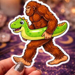 May include: A sticker featuring a cartoon illustration of a Bigfoot character carrying a green inflatable dinosaur pool float. The Bigfoot is brown and furry, with a serious expression. The float is green and yellow, with a smiling face.