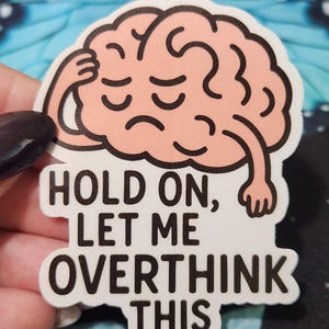 May include: A sticker featuring a cartoon brain in a light peach color, with closed eyes and a hand on its head. The sticker has the text "HOLD ON, LET ME OVERTHINK THIS" in black, bold letters.