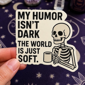 May include: A white sticker with the text "MY HUMOR ISN'T DARK THE WORLD IS JUST SOFT." features a black and white skeleton holding a mug. The sticker is held by a hand against a purple background.