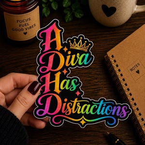 May include: A colorful sticker with the text "A Diva Has Distractions" in a rainbow gradient font, adorned with a gold crown. The sticker is held by a hand with dark nail polish, next to a lit candle and a coffee mug.