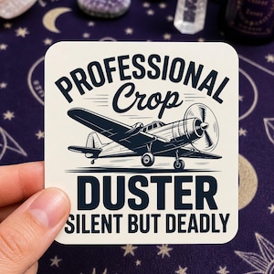 May include: A square sticker with a vintage airplane illustration. The sticker reads "PROFESSIONAL Crop DUSTER SILENT BUT DEADLY" in bold, dark blue text. The airplane is a dark blue silhouette.