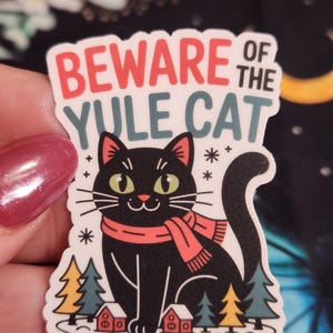 May include: A sticker featuring a black cat with green eyes and a red scarf, set against a snowy landscape with small houses and trees. The text "BEWARE OF THE YULE CAT" is displayed in red and teal.