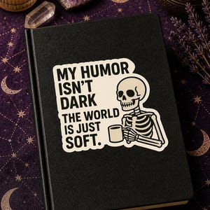 May include: Black journal with a sticker saying "MY HUMOR ISN'T DARK THE WORLD IS JUST SOFT." The sticker shows a skeleton with a mug. The journal rests on a purple surface with celestial patterns.