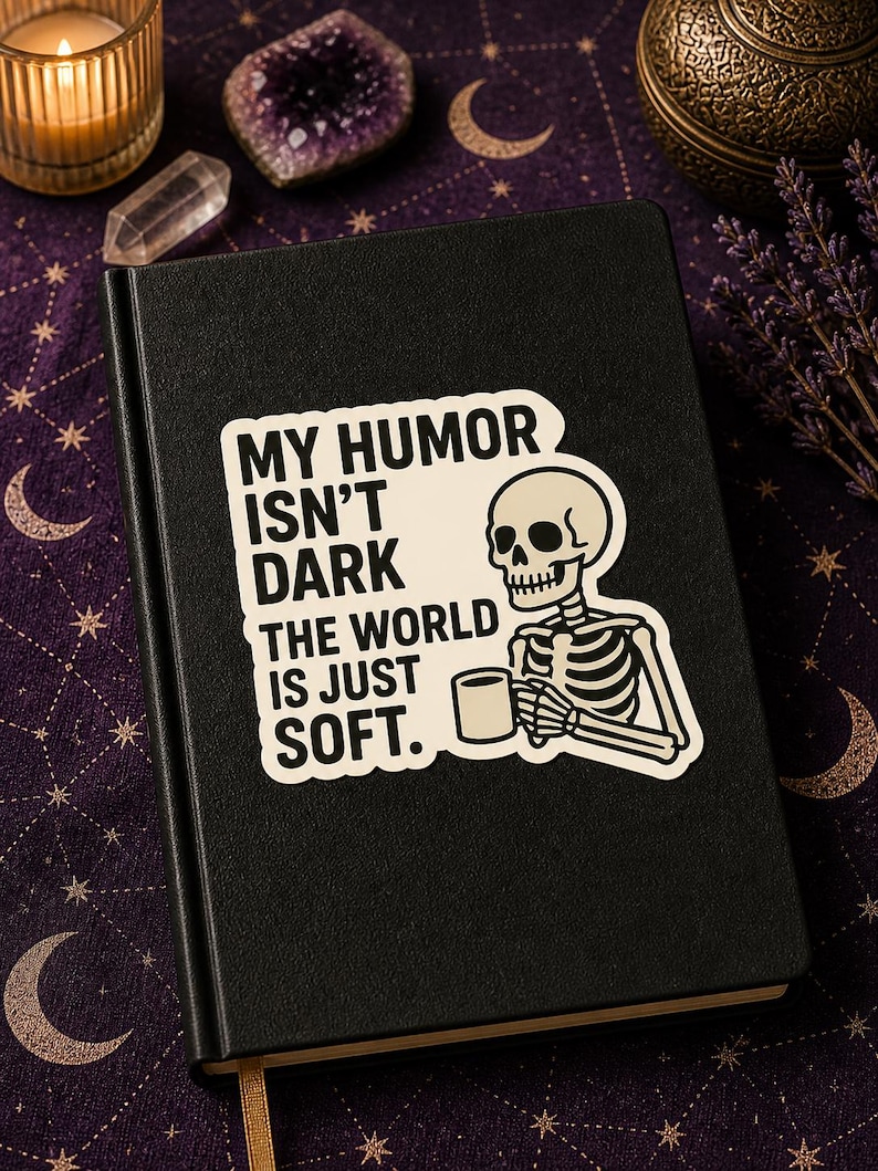 May include: Black journal with a sticker saying "MY HUMOR ISN'T DARK THE WORLD IS JUST SOFT." The sticker shows a skeleton with a mug. The journal rests on a purple surface with celestial patterns.
