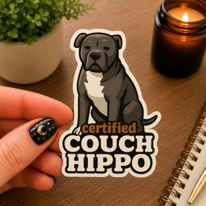 May include: A die-cut sticker featuring a dark gray and white pit bull dog illustration. Below the dog, the text reads "certified COUCH HIPPO" in bold, brown and white lettering. The sticker has a white border and a matte finish.