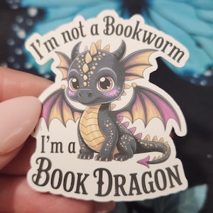 May include: A sticker featuring a cartoon dragon with the text "I'm not a Bookworm, I'm a Book Dragon." The dragon is black with purple and yellow wings, and has large, expressive eyes. The sticker is white with a glossy finish.