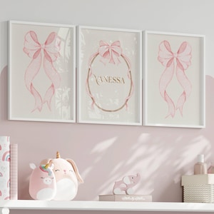 Peut inclure: Trois impressions encadrées avec des motifs de nœuds roses à l'aquarelle. L'impression centrale présente le nom "Vanessa" en lettres dorées dans un cadre ovale doré. Les impressions sont affichées au-dessus d'une étagère avec des objets décoratifs.