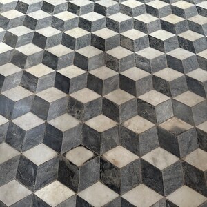 Może przedstawiać: Powtarzający się wzór szarych, czarnych i białych płytek tworzy iluzję optyczną trójwymiarowych kostek. Płytki ułożone są w siatkę, a każda kostka wydaje się wystawać z powierzchni. Całkowity efekt to wizualnie uderzający i dynamiczny design.