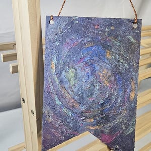 May include: A small, rectangular, purple, blue, and gold banner with a swirling, galaxy-like design. The banner is hanging from a string.