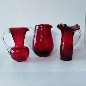 May include: Three vintage red glass jugs with clear handles. The jugs vary in shape, with one having a bulbous body, another a more cylindrical form, and the third with a flared rim. A classic glassware set.