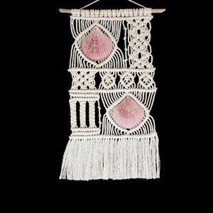 May include: A white macrame wall hanging with two pink seashells. The macrame is in a geometric pattern with fringe at the bottom.