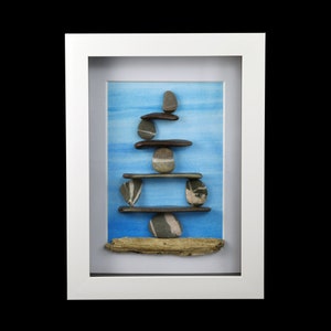 May include: A framed artwork featuring a rock balancing sculpture. The sculpture is made of various grey, brown, and white stones stacked on top of each other. The background is a blue watercolour wash. The sculpture is resting on a piece of driftwood.