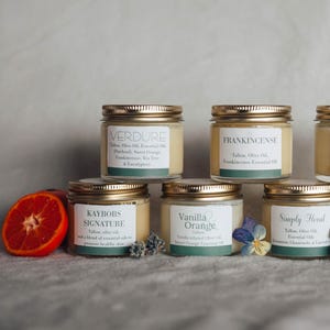 May include: A collection of eight small glass jars with gold lids, each containing a different scented body butter. The jars are arranged in two rows of four, with the labels facing the viewer. The labels include the names "Verdure", "Frankincense", "Leather Pine", "Kaybobs Signature", "Vanilla Orange", "Simply Floral", "Unscented Tallow & Olive Oil that's it!"