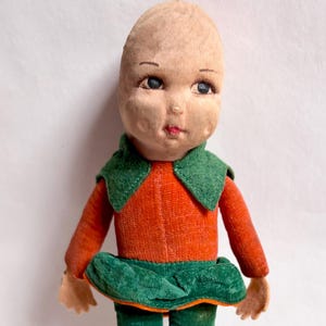 Puede incluir: Muñeca vintage con cabeza de tela y rasgos faciales pintados. La muñeca viste un top naranja con cuello verde, una falda verde y medias verdes. Los brazos y las manos de la muñeca son de tela color beige.