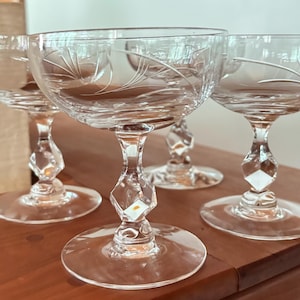 Op de afbeelding: Een set van vier heldere glazen coupes met een ondiepe kom en verfijnde, geëtste waaiervormige patronen. Elk glas heeft een gefacetteerde, ruitvormige steel en een ronde voet, wat zorgt voor een gestructureerde uitstraling.