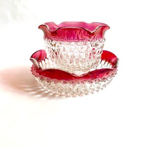 May include: A clear glass bowl and matching saucer with a ruby red rim. The bowl and saucer have a diamond pattern. The rim of the bowl and saucer have a ruffled edge. The set is on a white background.