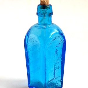May include: A vintage, rectangular, turquoise-blue glass bottle with a cork stopper. The bottle has embossed text and a rounded shoulder. The bottle is empty and sits against a white background.
