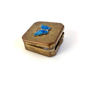 Puede incluir: Pequeña caja de latón antiguo con tapa con bisagras. La caja presenta un diseño de mariposa azul en la parte superior. La caja es rectangular con esquinas redondeadas y tiene un acabado deslustrado.