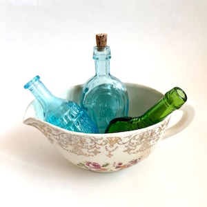 May include: A decorative white porcelain gravy boat with a floral and gold pattern, holding three glass bottles. Two bottles are blue, and one is green. One blue bottle has a cork stopper.