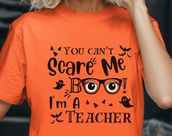 May include: Orange t-shirt with the text "You can't scare me, Boo! I'm a Teacher" in black. The design includes glasses, bats, and ghosts, perfect for Halloween or a teacher's wardrobe.
