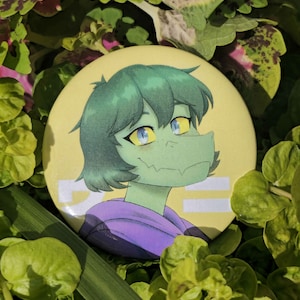 May include: A round, yellow button featuring a cartoon character with green skin, hair, and a purple hoodie. The character has yellow eyes and a reptilian snout. White Japanese text is visible on the button. The button is surrounded by green foliage.