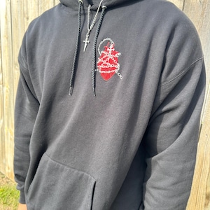 Puede incluir: Sudadera con capucha negra con bolsillo delantero y dise&ntilde;o de coraz&oacute;n rojo bordado con cadenas. Un collar de cadena plateada con un colgante de cruz cuelga de la capucha. La sudadera es de material suave.