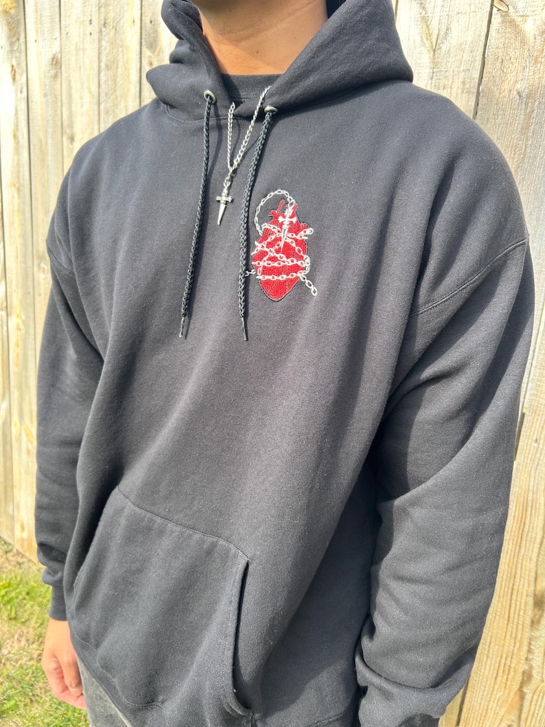 Puede incluir: Sudadera con capucha negra con bolsillo delantero y dise&ntilde;o de coraz&oacute;n rojo bordado con cadenas. Un collar de cadena plateada con un colgante de cruz cuelga de la capucha. La sudadera es de material suave.