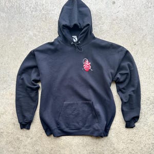 Puede incluir: Una sudadera con capucha negra con un bolsillo delantero y una capucha con cord&oacute;n. La sudadera presenta un dise&ntilde;o bordado en rojo y blanco en la parte superior izquierda del pecho. La prenda est&aacute; hecha de un material suave.