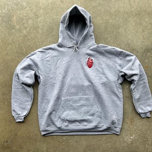 Puede incluir: Una sudadera con capucha gris con un bolsillo delantero y una capucha con cord&oacute;n. Un gr&aacute;fico rojo y blanco est&aacute; en el pecho izquierdo. La sudadera est&aacute; hecha de un material suave y c&oacute;modo.