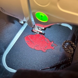 Puede incluir: Primer plano de una m&aacute;quina de bordar en funcionamiento. La m&aacute;quina est&aacute; bordando un dise&ntilde;o rojo sobre una tela negra. El dise&ntilde;o parece ser una forma estilizada de coraz&oacute;n o llama. Se ven la aguja y el prensatelas de la m&aacute;quina.