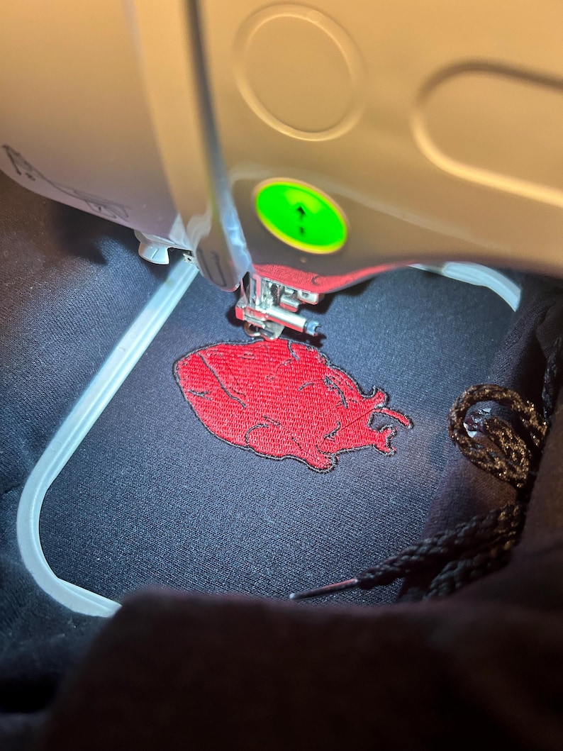 Puede incluir: Primer plano de una m&aacute;quina de bordar en funcionamiento. La m&aacute;quina est&aacute; bordando un dise&ntilde;o rojo sobre una tela negra. El dise&ntilde;o parece ser una forma estilizada de coraz&oacute;n o llama. Se ven la aguja y el prensatelas de la m&aacute;quina.