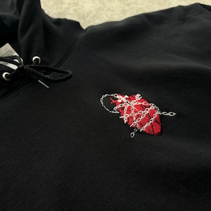Puede incluir: Sudadera con capucha negra con cord&oacute;n y un dise&ntilde;o de coraz&oacute;n rojo bordado envuelto en una cadena plateada. La sudadera con capucha est&aacute; hecha de un material suave y tiene un estilo cl&aacute;sico e informal.