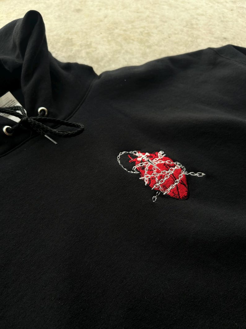 Puede incluir: Sudadera con capucha negra con cord&oacute;n y un dise&ntilde;o de coraz&oacute;n rojo bordado envuelto en una cadena plateada. La sudadera con capucha est&aacute; hecha de un material suave y tiene un estilo cl&aacute;sico e informal.