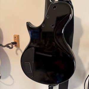 Könnte beinhalten: Eine glänzend schwarze E-Bassgitarre, von hinten betrachtet, an einer Wand befestigt. Das Instrument hat einen konturierten Korpus, sichtbare Stimmwirbel und einen schwarzen Gurt. Die Bassgitarre wird von einer Wandhalterung gehalten.