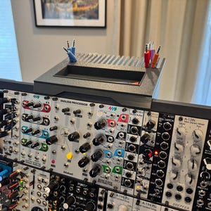 May include: A modular synthesizer system with various modules, knobs, and jacks. A black tray with slots and colored cables sits atop the system. The modules have labels like "Endless Processor" and "Quad Buffer".