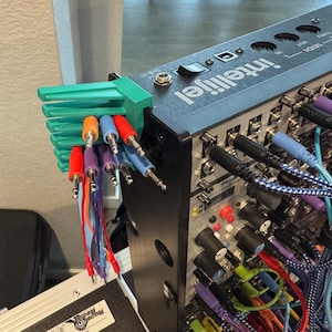 Puede incluir: Un sintetizador modular con un organizador de cables verde que sostiene cables de audio multicolores. El sintetizador tiene una carcasa de metal negro con la marca "Intellijel" impresa en la parte superior.