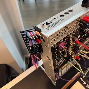 May include: A silver Intellijel Eurorack modular synthesizer with numerous jacks, knobs, and colorful patch cables. A black cable organizer holds the patch cables. The synthesizer is on a black surface.