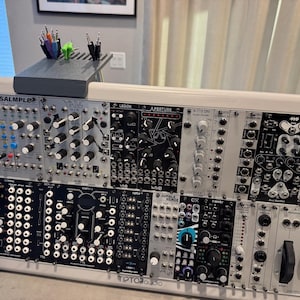 May include: Modular synthesizer with multiple modules, knobs, and jacks. The modules are labeled with text such as "SQUID SALMPLE", "XNYS", and "LEGION". A small gray shelf holds pens and cables.