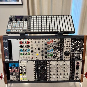 May include: A modular synthesizer setup featuring a black and silver controller on top of a wooden rack. The rack houses various electronic modules with knobs, buttons, and displays. The setup includes the text "OR INSTRO" and "ONE".