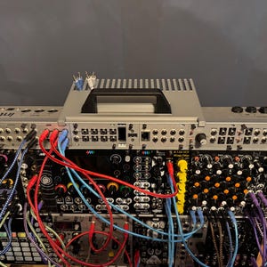 May include: A close-up view of a modular synthesizer with a variety of modules and patch cables. The synthesizer is connected to a mixer with a silver faceplate and a black screen. The mixer has a handle on the top and is labeled "Intellijel".
