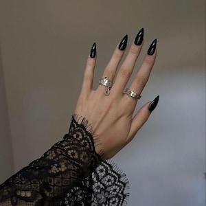 May include: Hand with long, black, pointed fingernails, adorned with two silver rings. One ring features a safety pin design, the other has a floral pattern. The wrist is partially covered by a black lace sleeve.